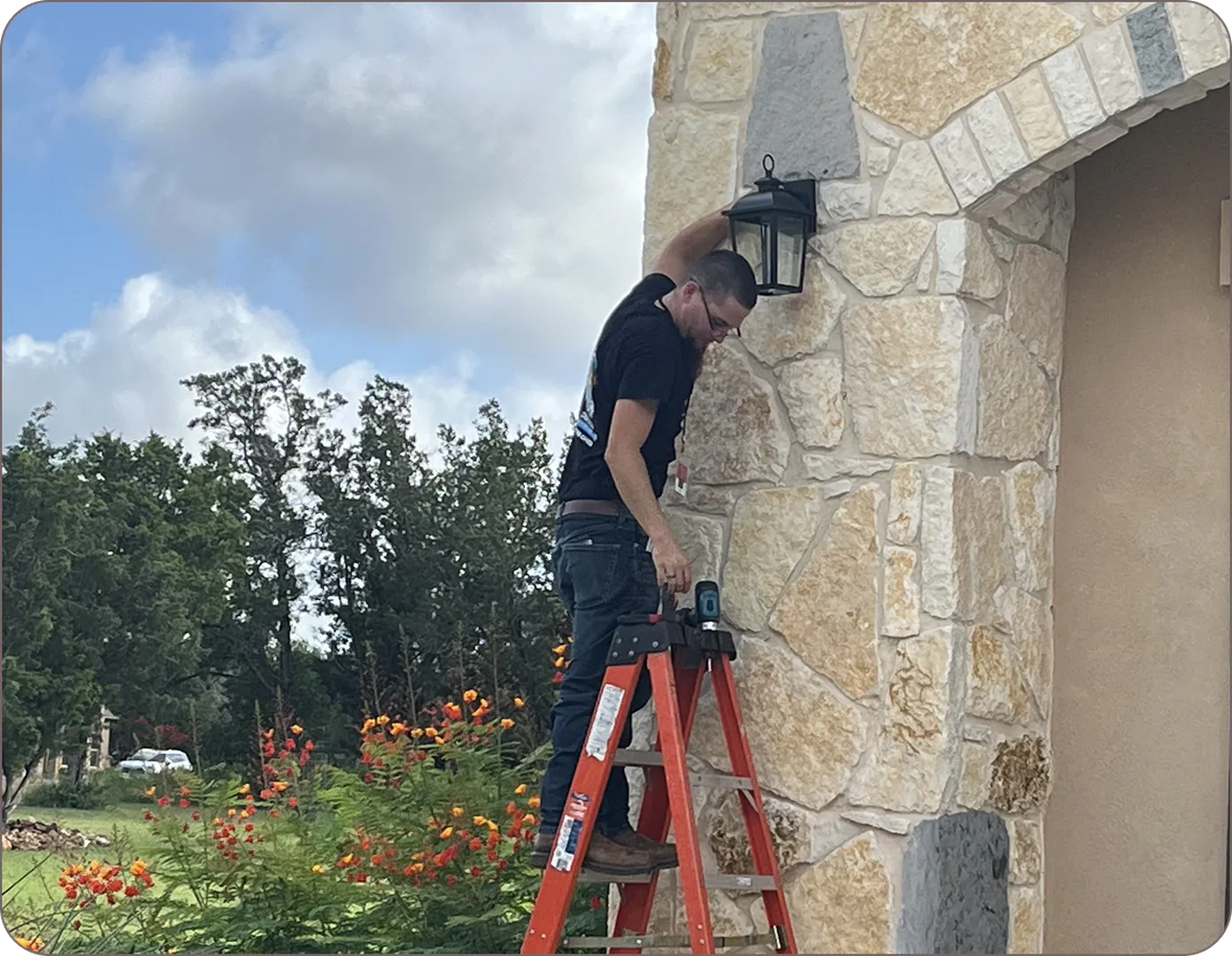 A man on a red ladder fixes a lantern on a stone wall with greenery and orange flowers.