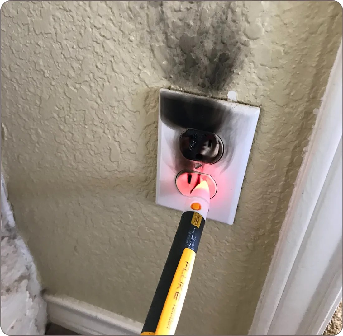 Burnt electrical outlet on a beige wall with soot; a red voltage tester highlights the issue.
