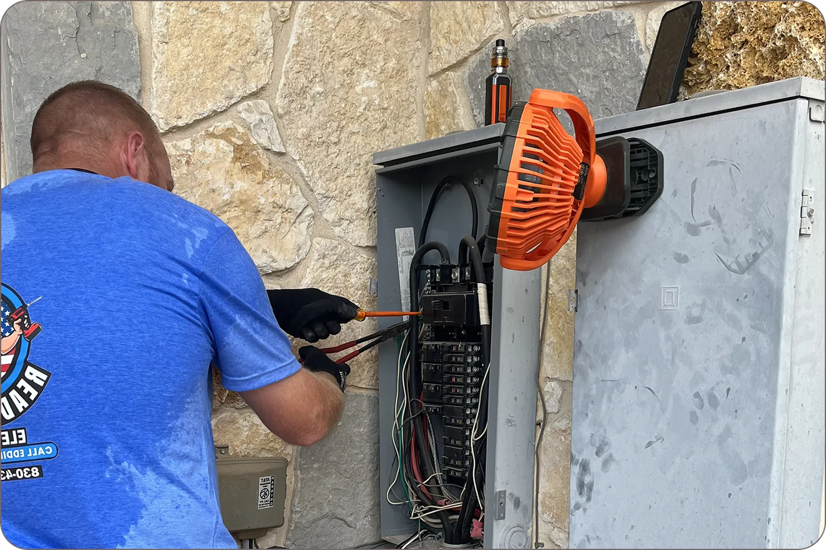 A man in a blue shirt and gloves is working on an electrical panel outside.