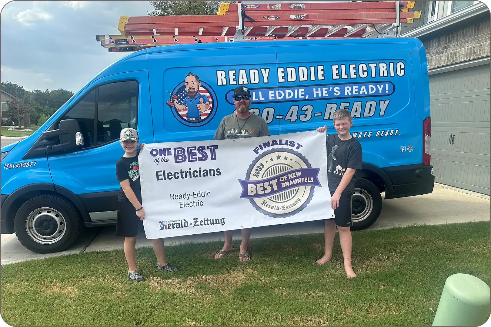 Eddie Burton and two boys hold a 'Best Electricians' banner in front of a blue Ready Eddie van.