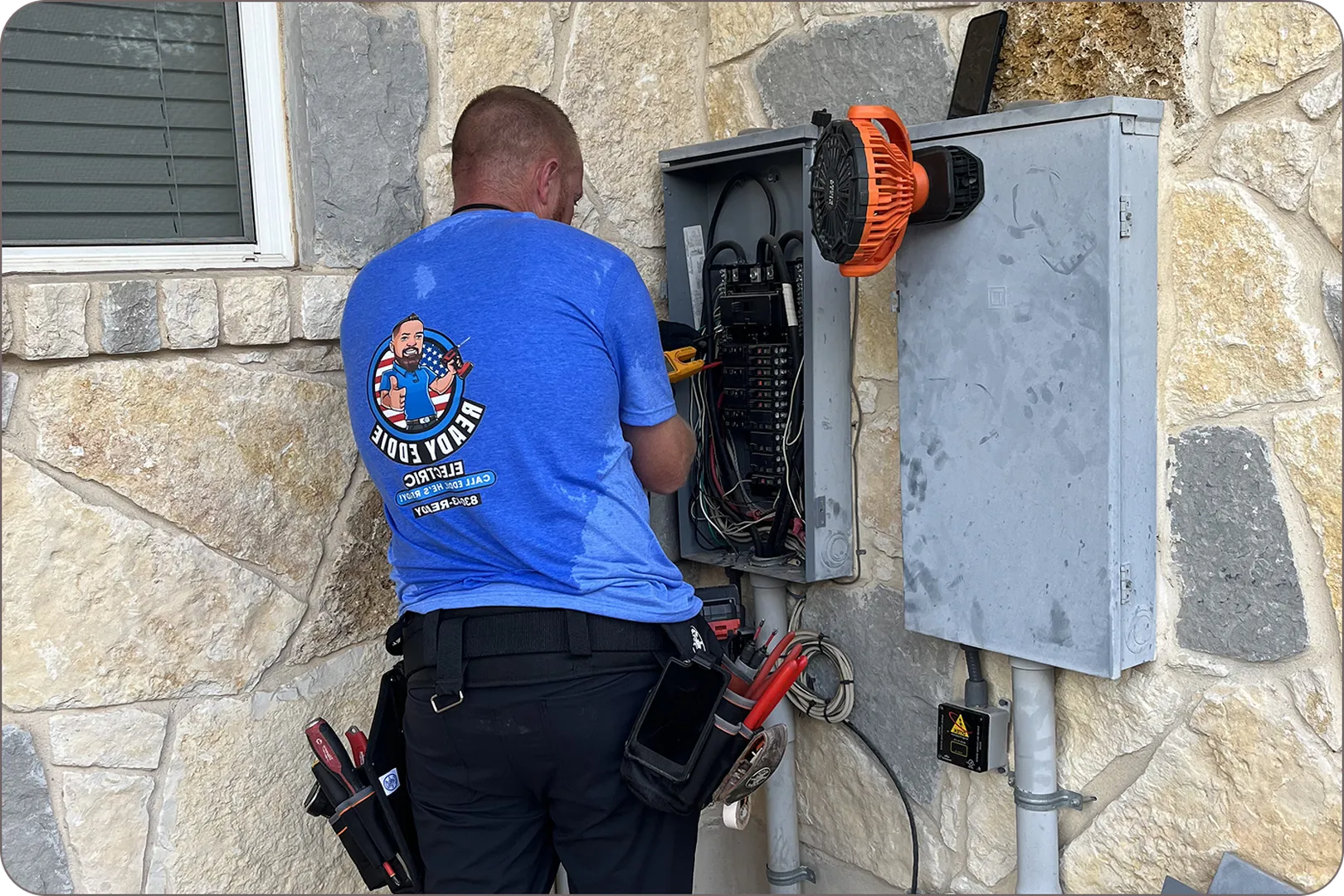 An electrician works on a panel on a stone wall with a utility belt.