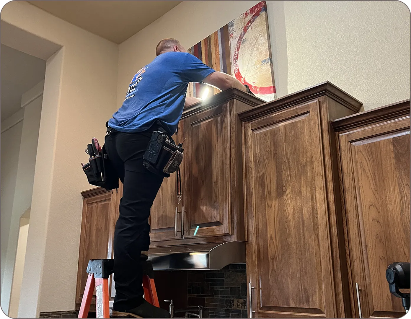 Eddie Burton using tools on his belt to fix the lighting in cabinets while perched on a ladder.