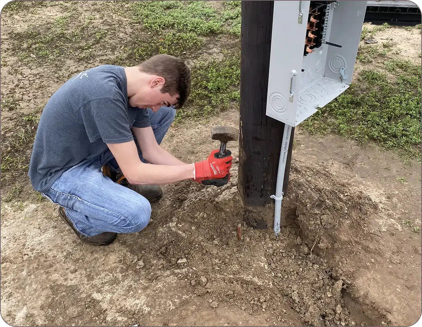 A man in jeans and a shirt is driving a rod into the ground at a wooden utility pole.