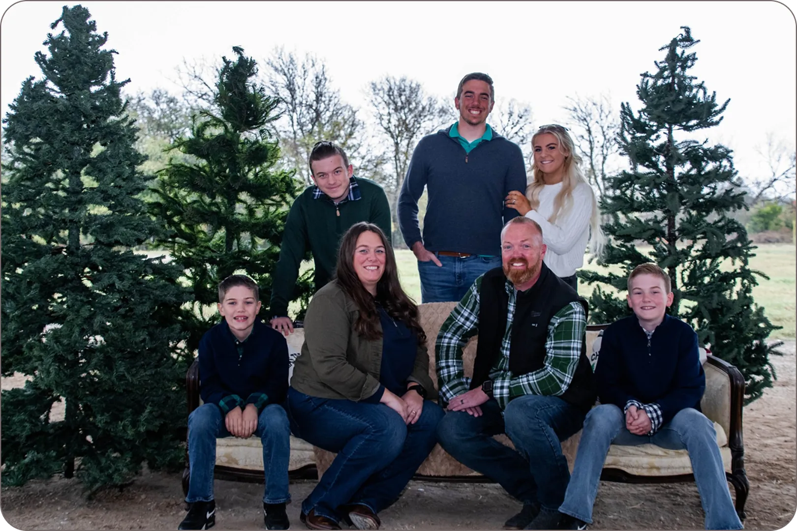 Eddie Burton and family smile on an outdoor sofa by pine trees.